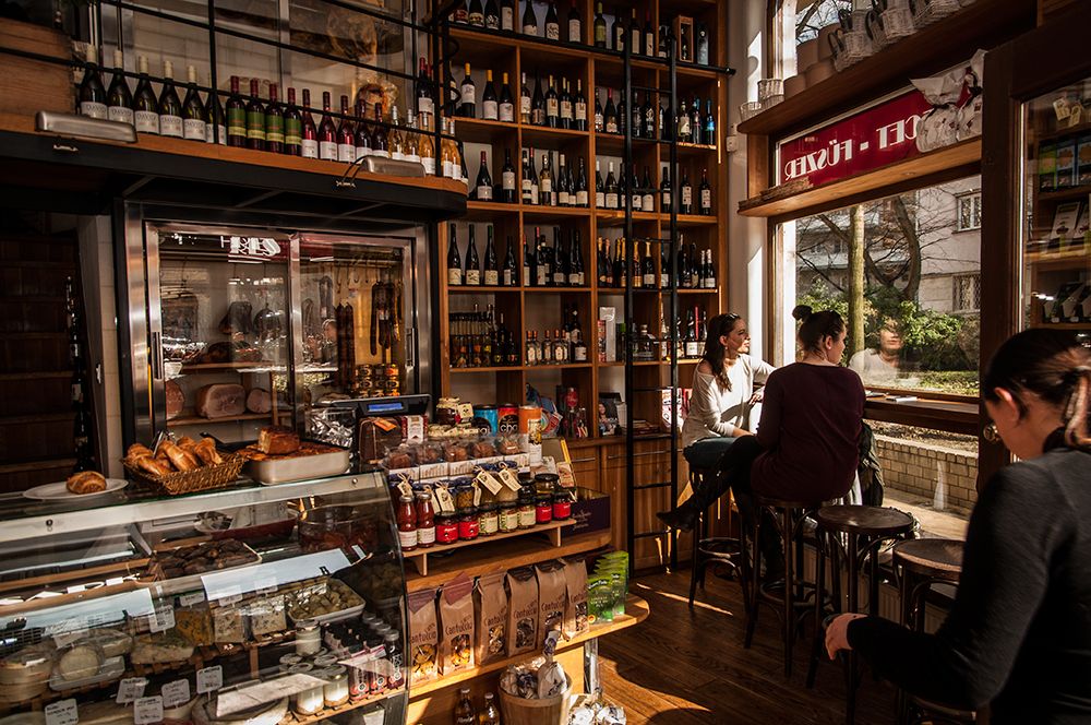 Gourmet shop interior with floor-to-ceiling wine shelves, glass deli case, and patrons at the bar.